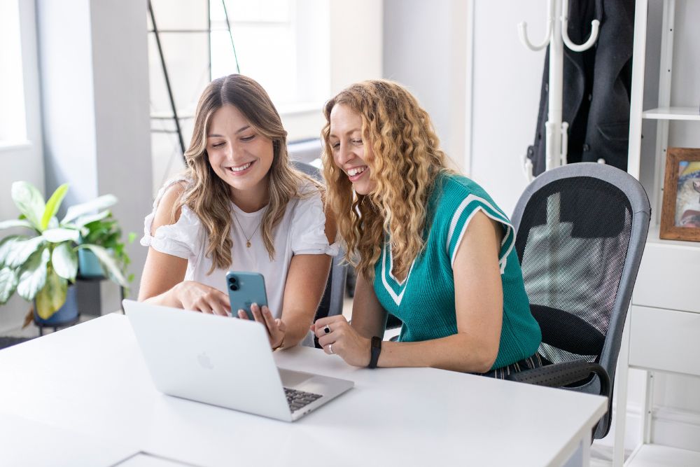 Two women smiling at a laptop in a brightly lit office